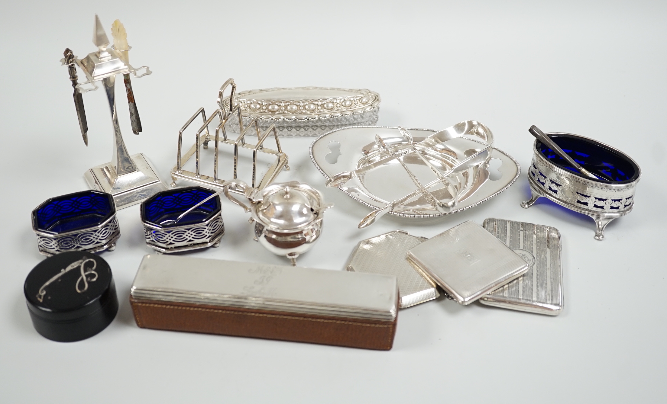 A group of mixed silver ware including a manicure stand, toastrack, purse, two compacts, four condiments, two pairs of sugar tongs, a stand and two mounted glass toilet jars.